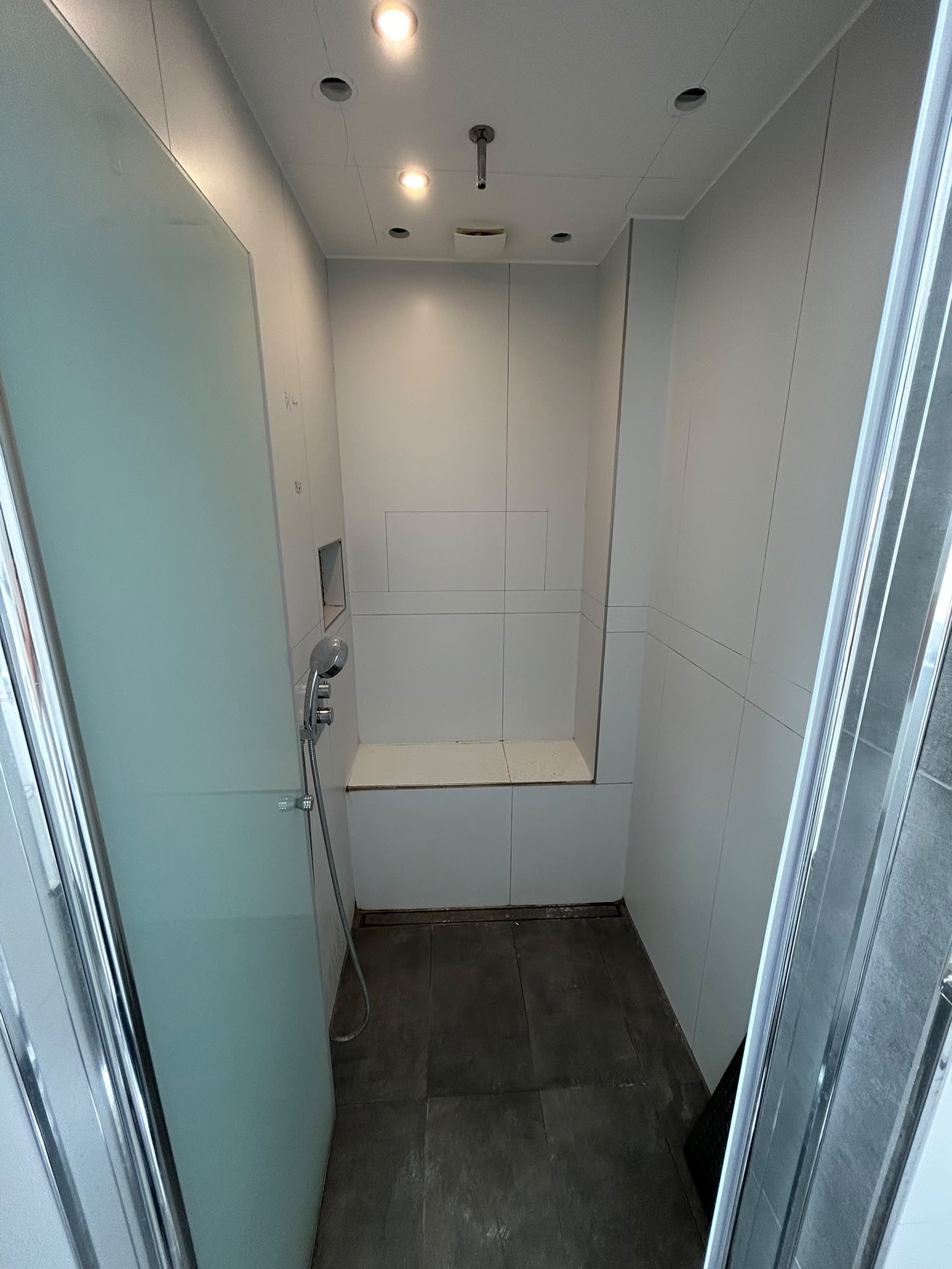 Dusche vor der Sanierung: weiß gefliest, mit Sitzbank und Duschrinne