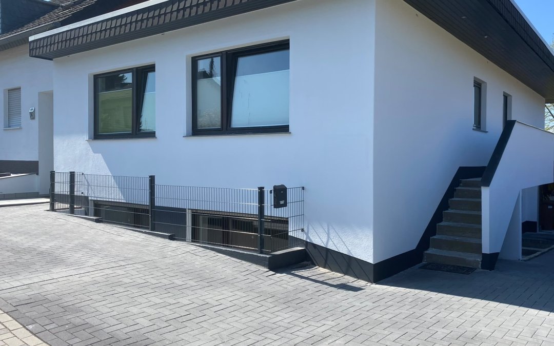 Dasselbe Haus nach der Sanierung: gedämmte Fassade mit hellem Putz, neue Fenster, Stabmattenzaun und neues Pflaster